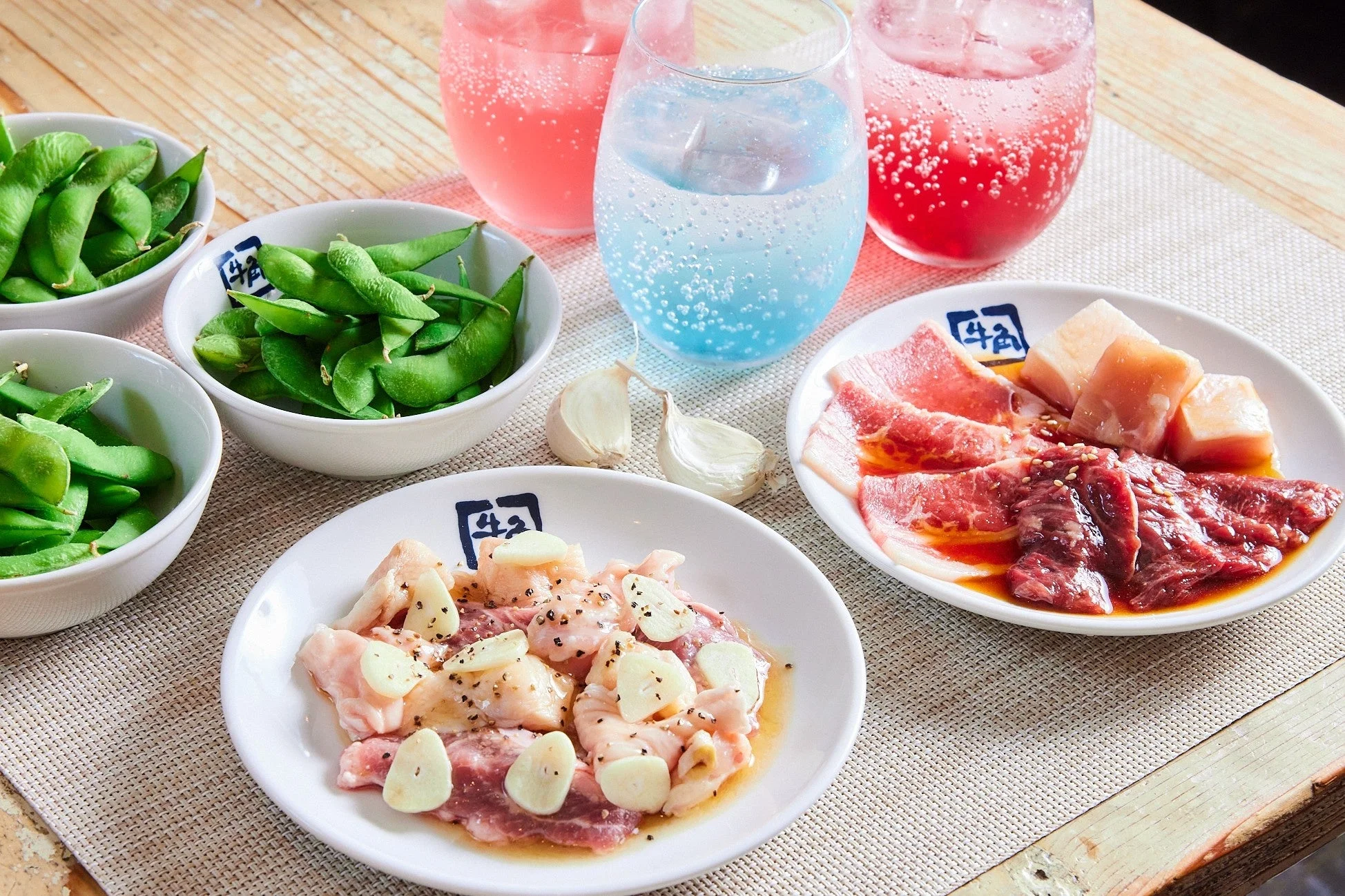 焼肉用の様々な生肉、枝豆、カラフルなソーダドリンクが並べられた食卓の様子。牛角のロゴが入った皿に盛られた肉には、ニンニクのスライスが添えられています。