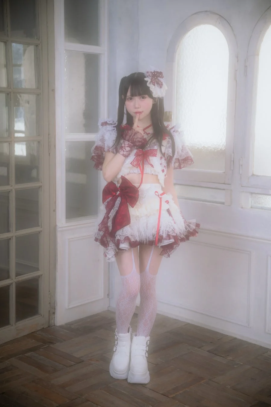 白と赤のフリルが特徴的なかわいらしい衣装をまとった女性が、室内でポーズをとっています。ツインテールの髪型と、指を唇に当てる仕草が印象的で、ドールのような雰囲気を醸し出しています。