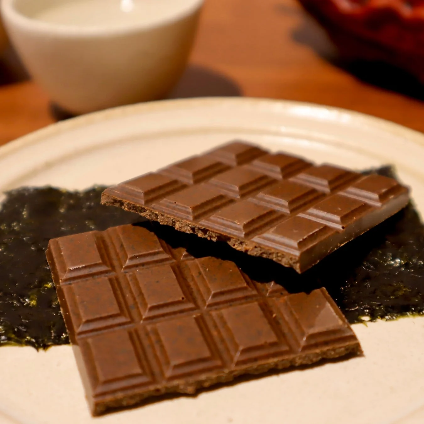 白い皿に置かれた2枚の板チョコレート。チョコレートの下には海苔のような緑色のペーストがあり、ユニークな組み合わせのデザートです。背景にはぼやけたカップが見えます。