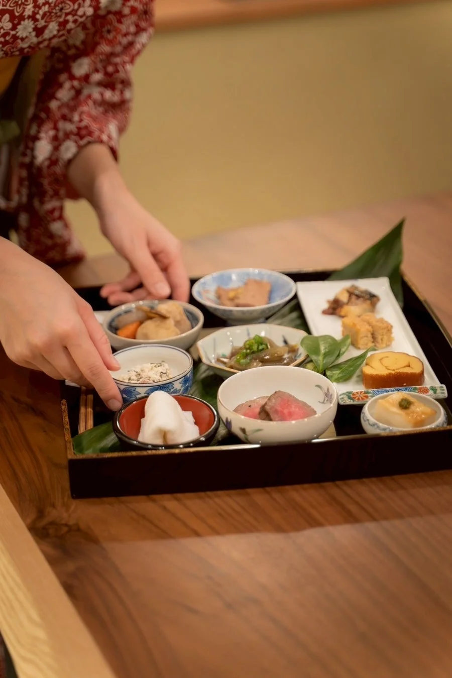 着物を着た人物が、木製のテーブルに置かれたお膳に並べられた美しい和食の小鉢料理を丁寧に整えている様子。様々な種類の料理が盛り付けられた、伝統的な日本の食事が写されています。