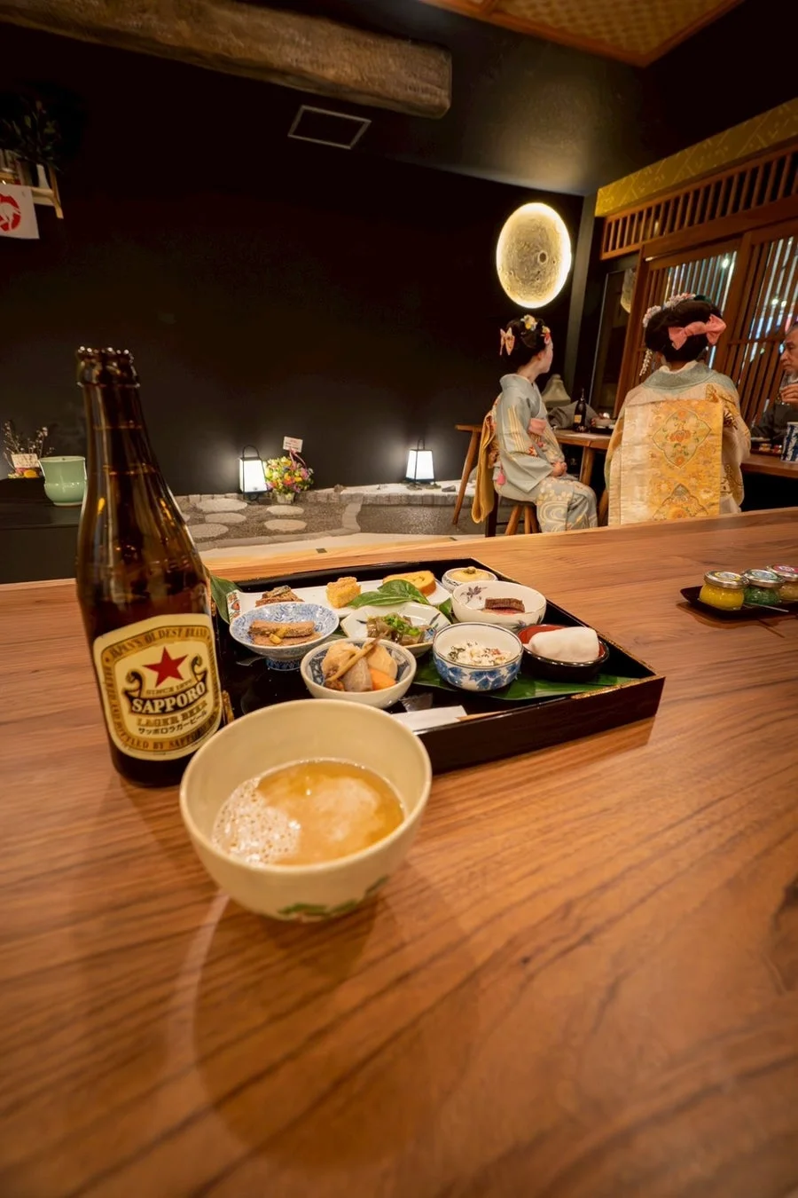 伝統的な雰囲気の店内で、着物を着た女性たちが食事を楽しんでいます。手前にはサッポロビールと小鉢が並べられた和食の膳があり、日本の食文化が感じられる一枚です。