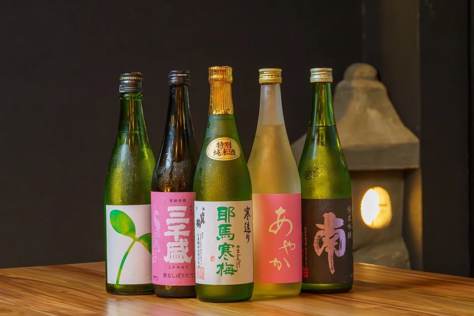 木のテーブルに並べられた5本の冷えた日本酒の瓶。結露が冷たさを伝え、背景の石灯籠が和の雰囲気を醸し出している。