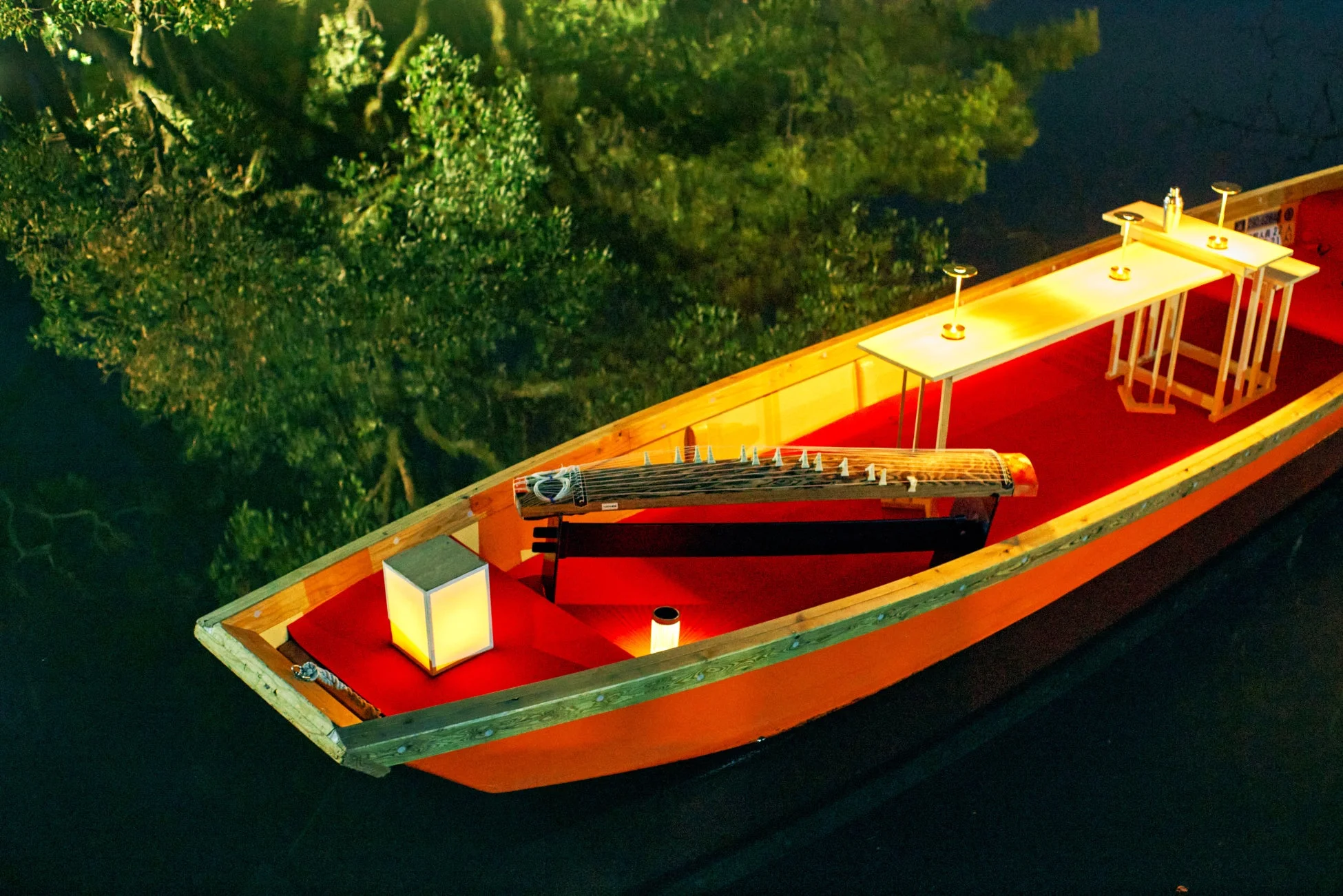 水面に浮かぶ船の内部には琴が置かれ、複数のテーブルには灯りが灯っています