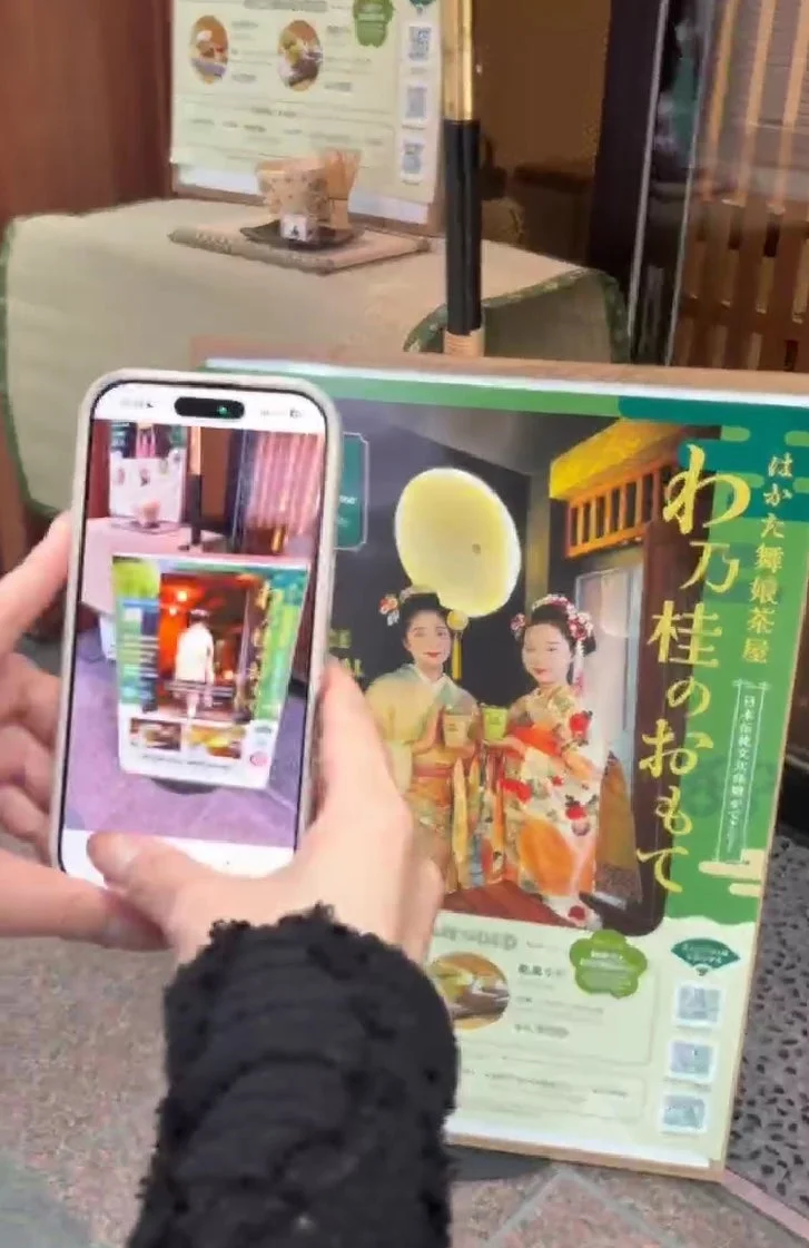スマートフォンで、和服姿の舞妓が描かれた「はかた舞妓茶屋」のポスターを撮影している。日本の伝統文化体験を宣伝する内容で、メニューらしきものも見える。