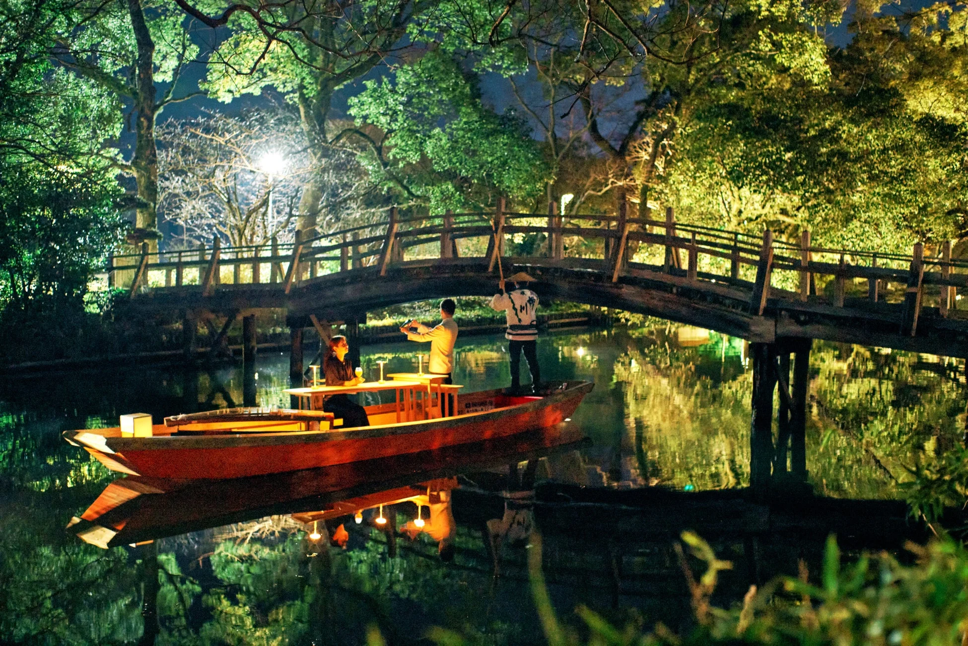 ライトアップされた木々が水面に映る中、木製の橋の下を小舟が進む夜の風景