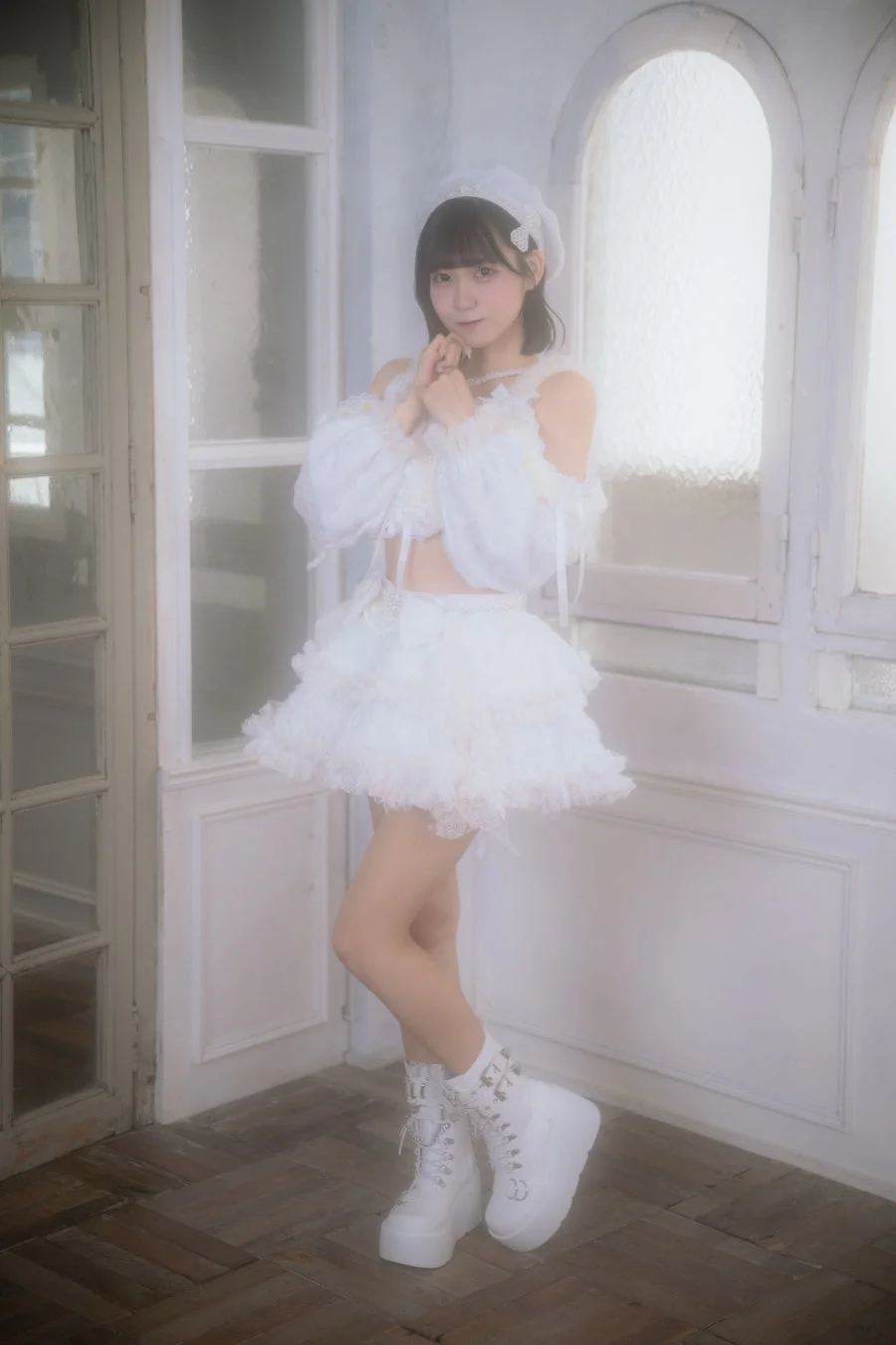 白いフリルとレースの衣装を身につけた若い女性が、白い窓を背景にポーズをとっています。ふんわりとした雰囲気で、厚底ブーツが特徴的です。