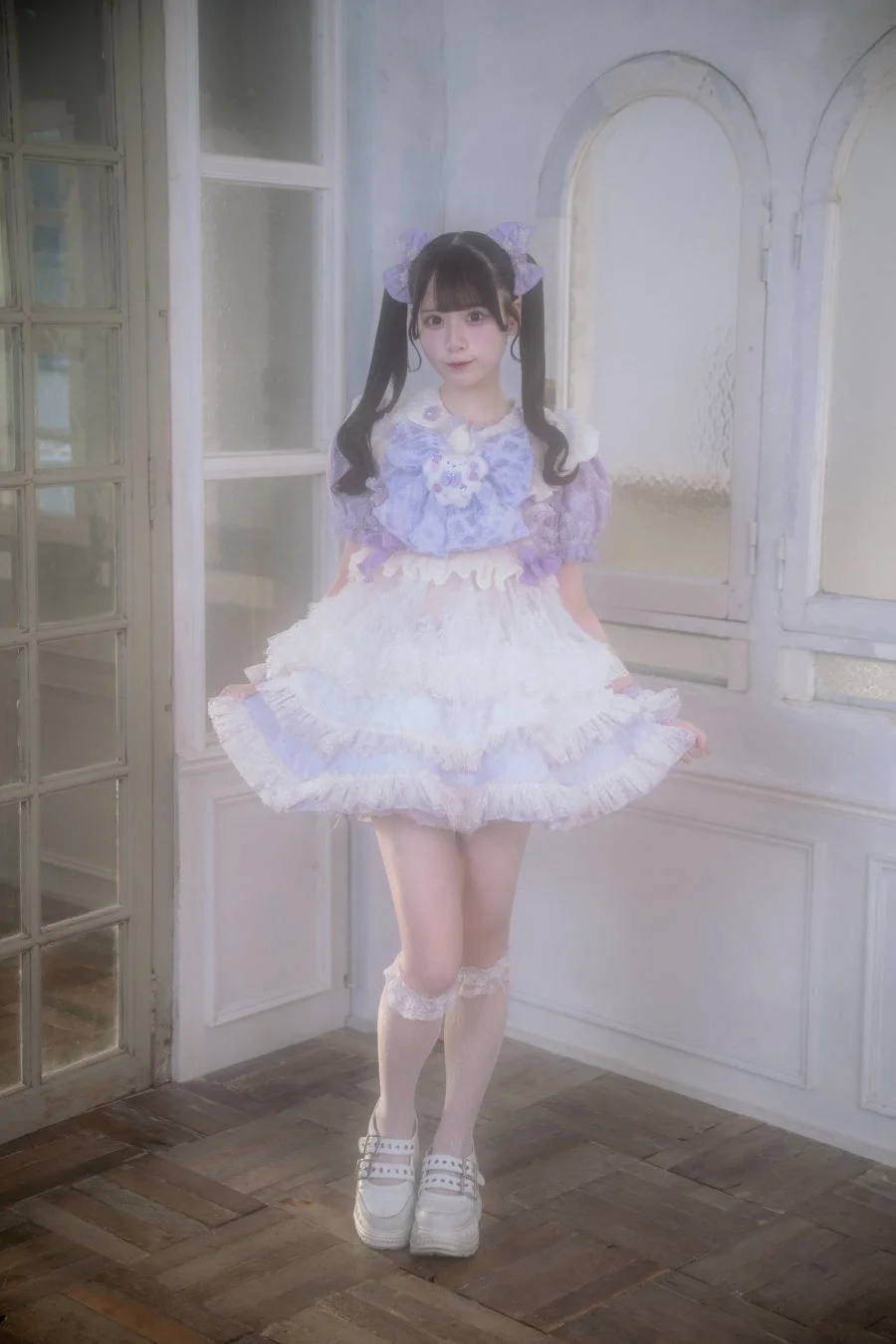 紫と白のフリルが特徴的なドレスを着用し、ツインテールにリボンを飾った女性の全身ポートレート。甘く可愛らしい雰囲気で、白い背景の室内でカメラを見つめている。