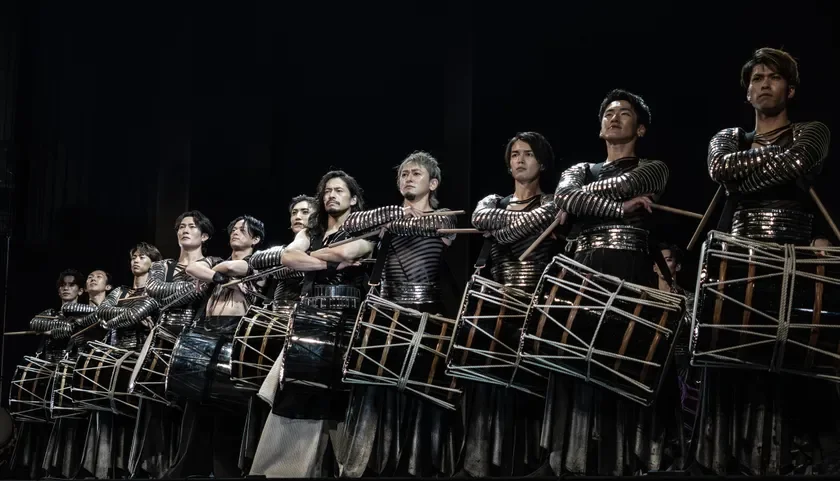 メタリックな衣装をまとった複数の男性が、和太鼓の前に立ち、ドラムスティックを構えている舞台でのパフォーマンスの様子です。