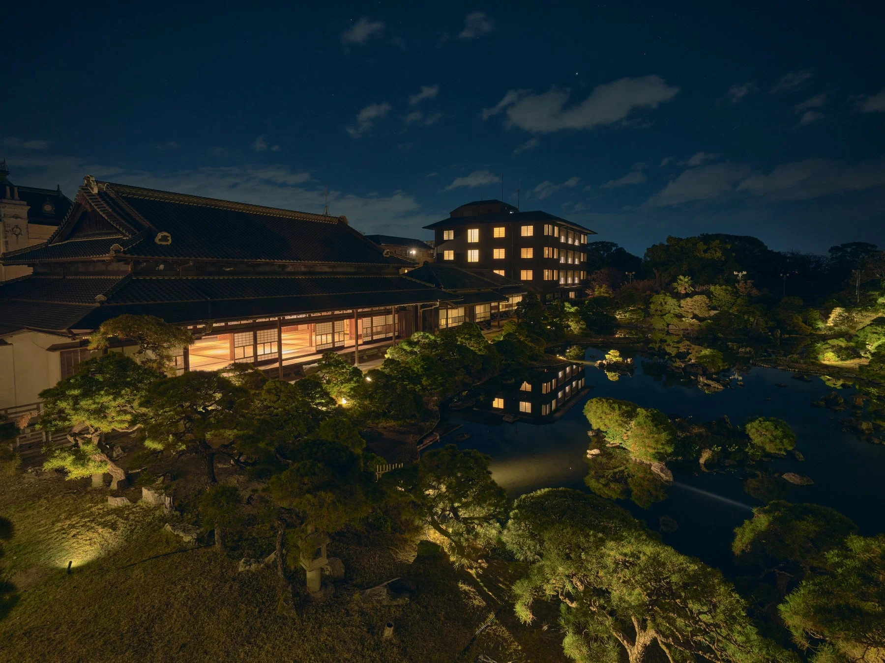 夜の日本庭園と伝統的な建築物が美しくライトアップされており、水面に光が反射している幻想的な風景