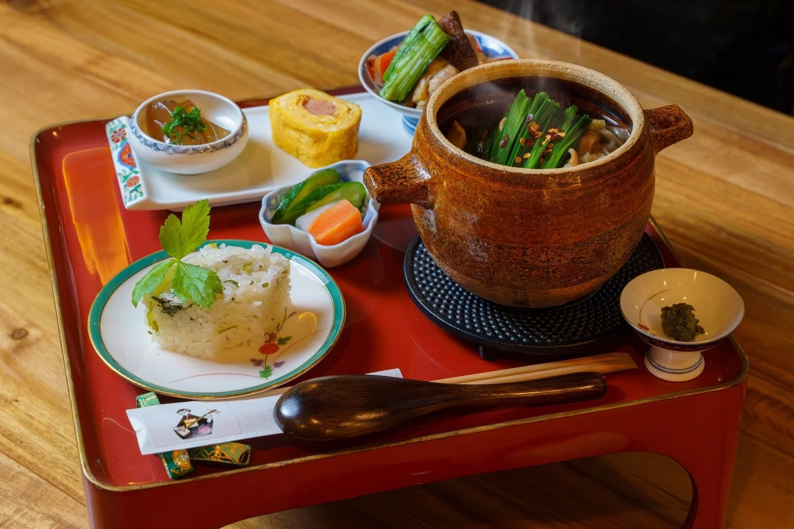 温かい湯気が立ち上る土鍋を中心に、ご飯、卵焼き、漬物など様々な和食の小鉢が赤いお膳に並べられた、彩り豊かで美味しそうな定食の風景です。