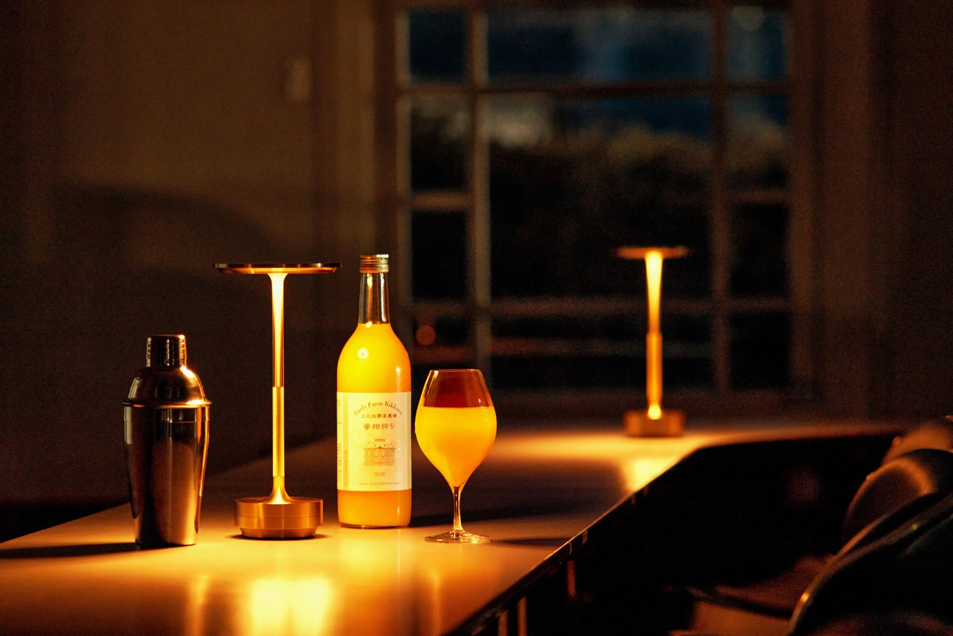 暖色の照明が印象的な空間に、オレンジ色の飲み物が入ったボトルとグラス、カクテルシェーカーが置かれている