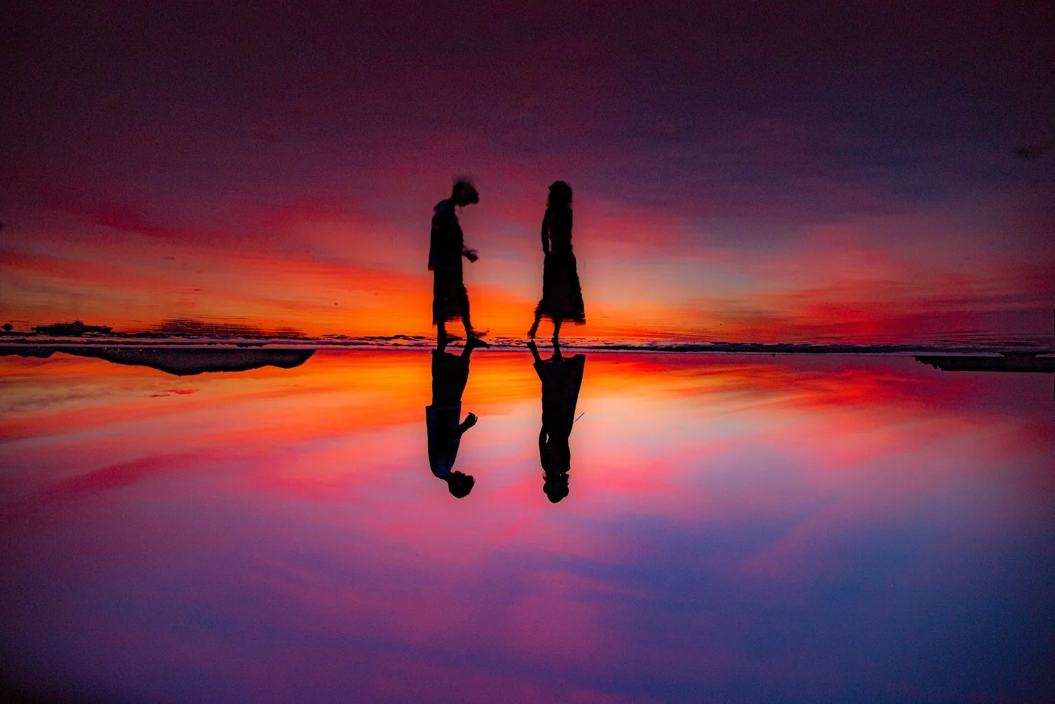 鮮やかな夕焼け空が水面に映り込み、幻想的な景色を作り出しています。二人の人物のシルエットが水面にもくっきりと反射し、オレンジ、赤、紫のグラデーションが広がる美しい瞬間を捉えた一枚です。