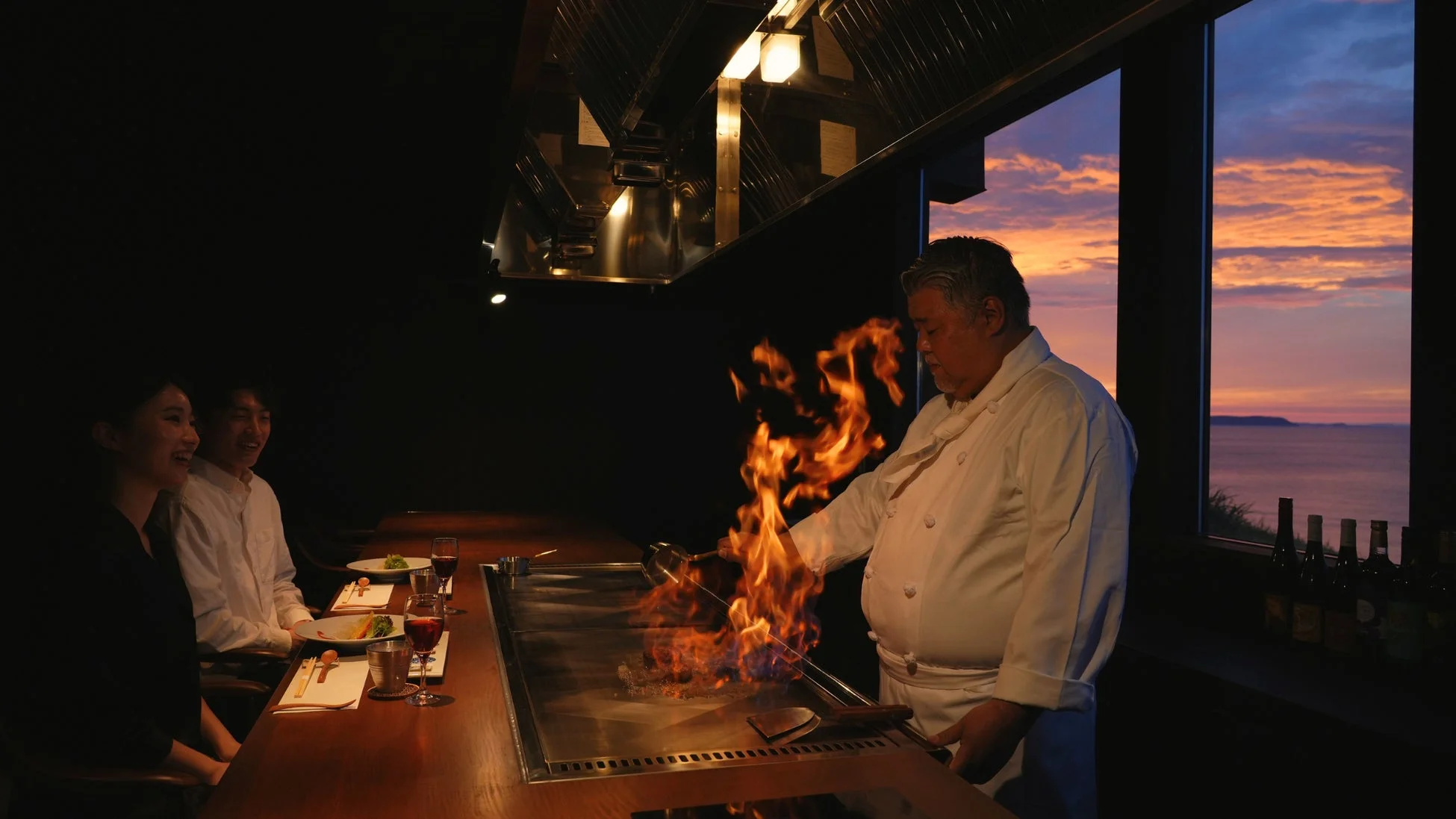 鉄板焼きレストランでシェフが炎を上げて調理しており、客がその様子を楽しんでいます
