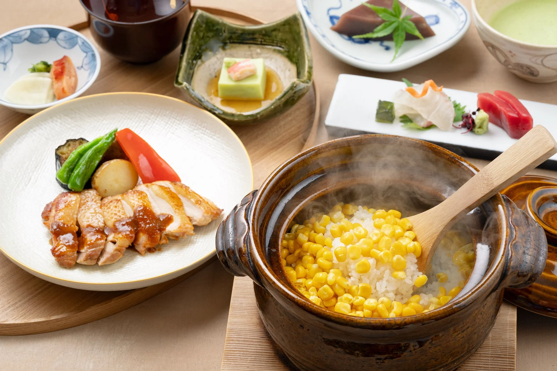 木製の膳に並べられた豪華な和食の定食です。照り焼きチキンと彩り野菜、湯気の立つとうもろこしご飯を中心に、刺身、和え物、汁物、デザートなど品数豊富でバランスの取れた食事が食欲をそそります