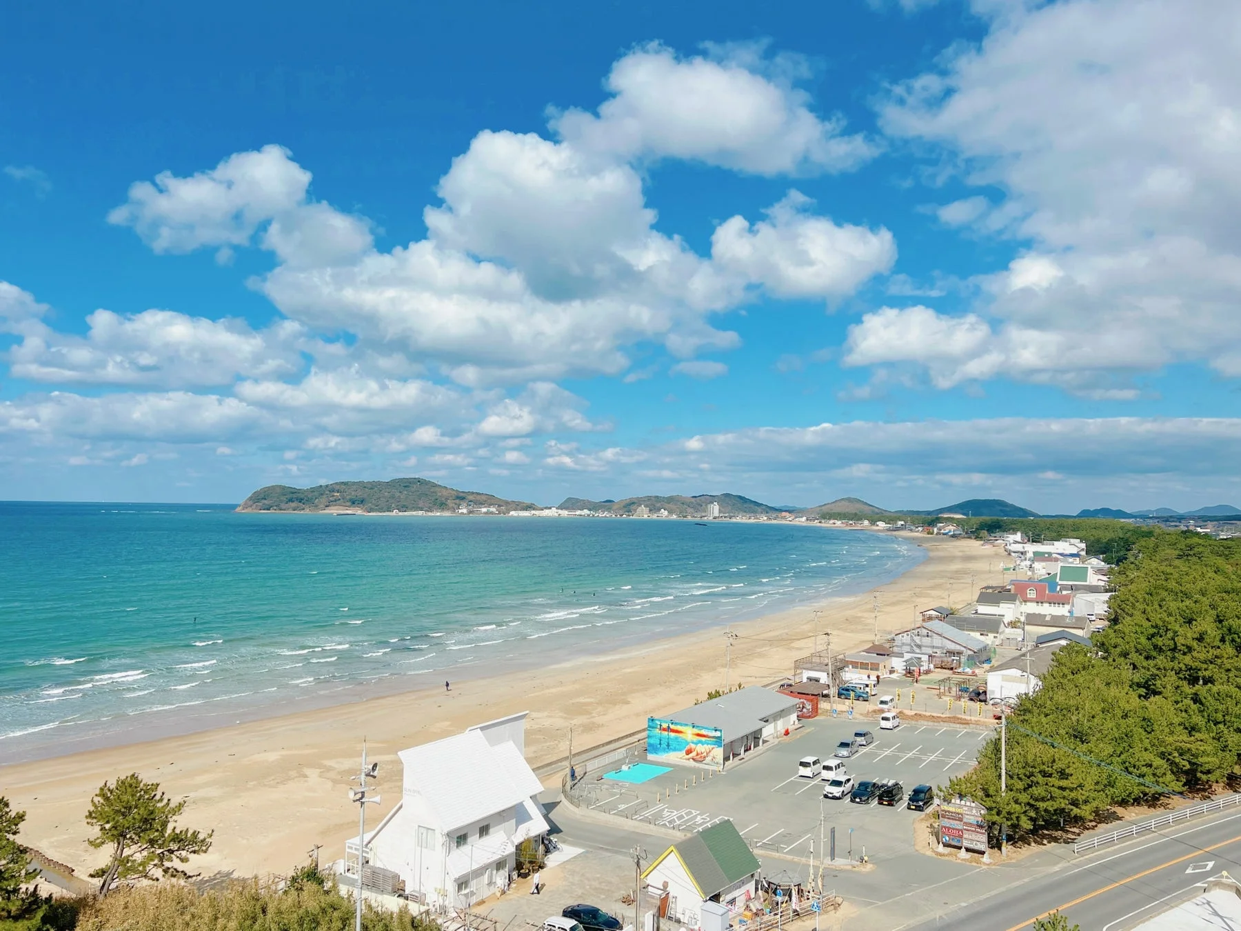 青い空と白い雲が広がる美しい海岸線の風景です。ターコイズブルーの海と広い砂浜が続き、沿岸には建物や駐車場、緑豊かな松の木が並んでいます。遠くには山々が見え、穏やかな波が打ち寄せる開放的な景色です。