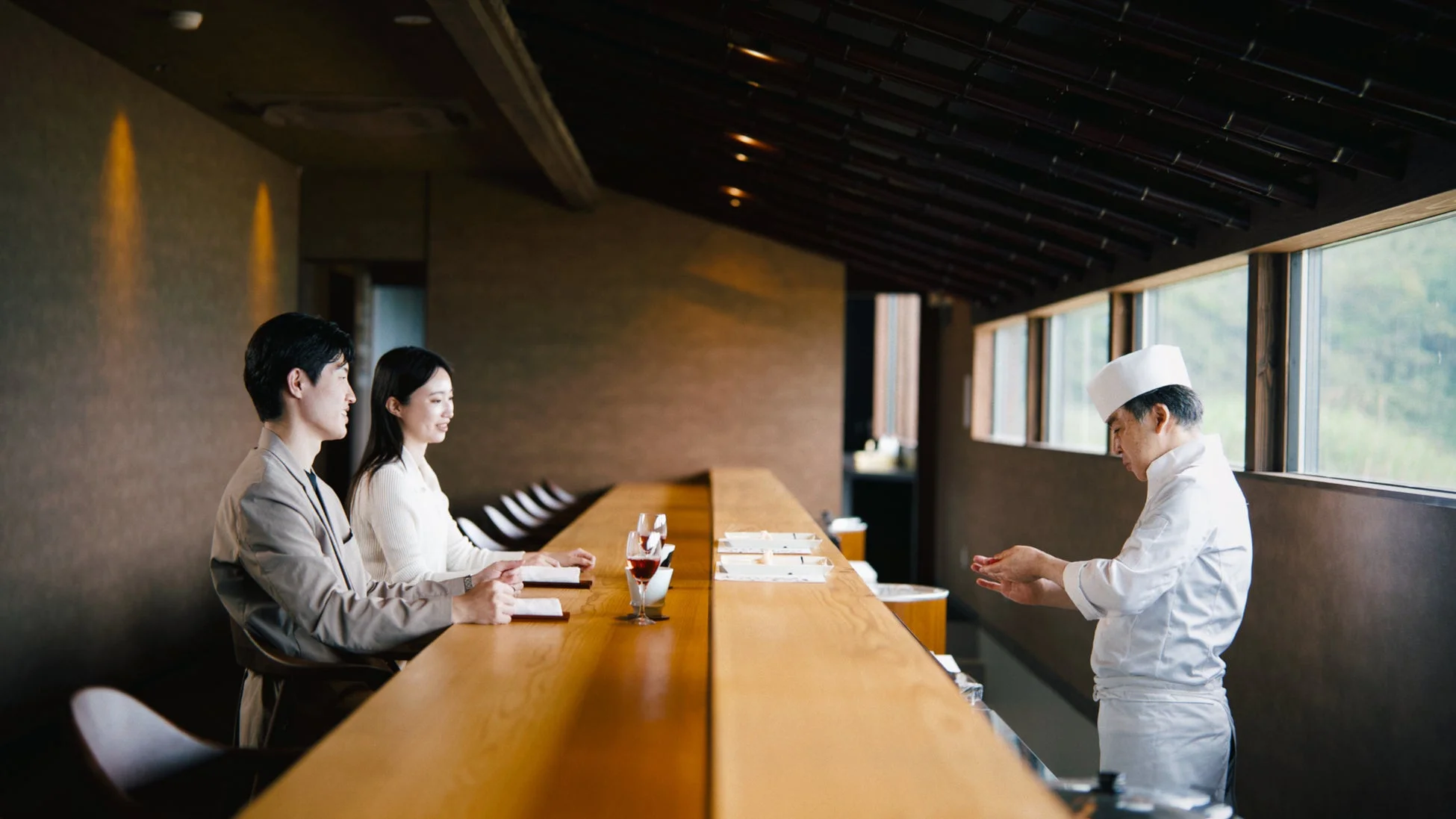 高級感のある日本料理店で、板前がカウンター席に座るカップルの前で寿司を握っている様子