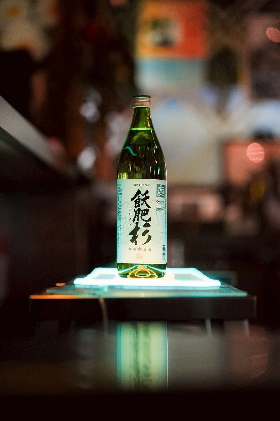 緑色のボトルに入った日本の本格焼酎「飫肥杉」が、ライトアップされた台座の上に置かれている写真です。背景はぼやけており、バーや居酒屋のような雰囲気が感じられます。