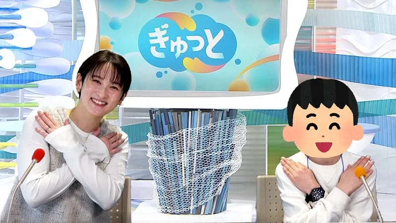 テレビスタジオで、ショートヘアの女性が笑顔で腕を組むポーズをしており、隣には顔がイラスト化された男性も同じポーズをしています。背景のモニターには「ぎゅっと」という文字が表示されています。