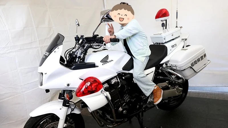 顔が隠された人物が、白いホンダ製の白バイに乗り、ピースサインをしている様子です。車両には「福岡県警察」の文字と赤色灯が見えます。