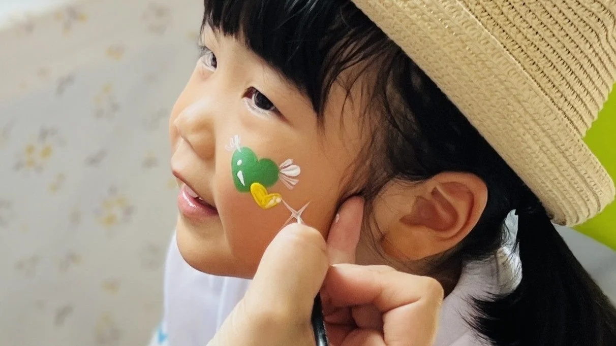 麦わら帽子をかぶった女の子が、頬に緑と黄色のハートマークのフェイスペイントを施してもらっている様子です。笑顔で楽しそうな表情をしています。