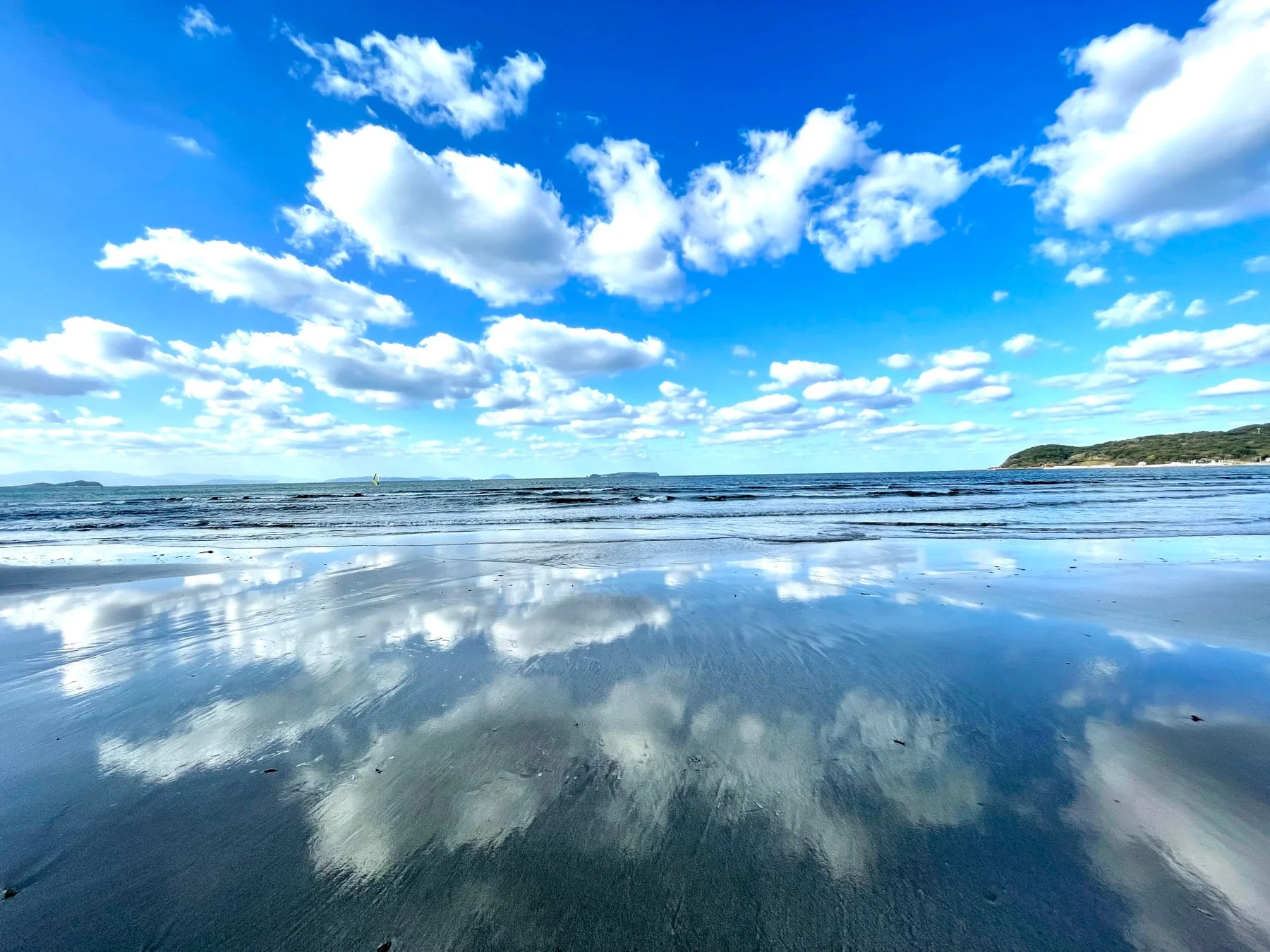 青い空に白い雲が浮かび、その雲が波打ち際の濡れた砂浜に美しく反射しているビーチの風景。遠くには穏やかな海と緑豊かな海岸線が見え、小さなヨットが浮かんでいる。