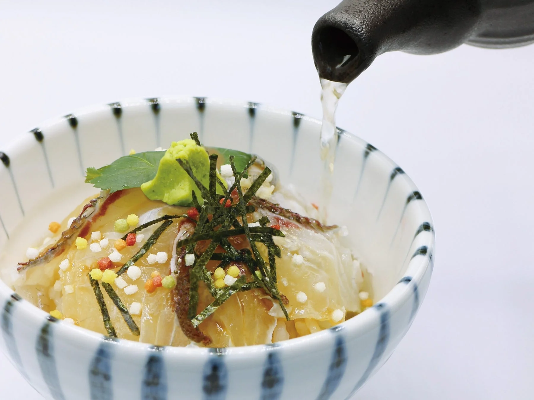 器に盛り付けられた鯛の切り身が乗ったご飯に、急須から温かい出汁またはお茶が注がれている和食の様子です。ワサビ、海苔、カラフルなあられがトッピングされています。