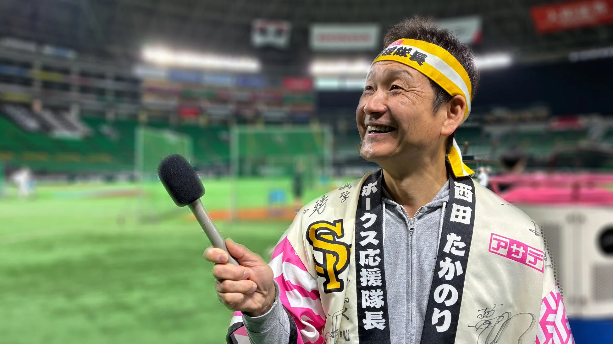 野球場で笑顔の男性がマイクを手にしている。彼は「応援隊長」と書かれたハチマキと、ホークスの応援隊長を示す文字や「西田たかのり」の名前が入った法被を着用しており、プロ野球の応援イベントの様子を捉えた一枚。