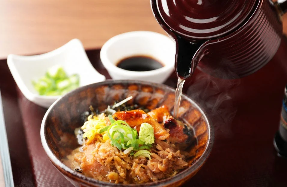 湯気を立てながら、だしを注ぎ入れる様子が食欲をそそる、うなぎのお茶漬け（ひつまぶし）の画像です。薬味としてネギ、ワサビ、錦糸卵が添えられています。