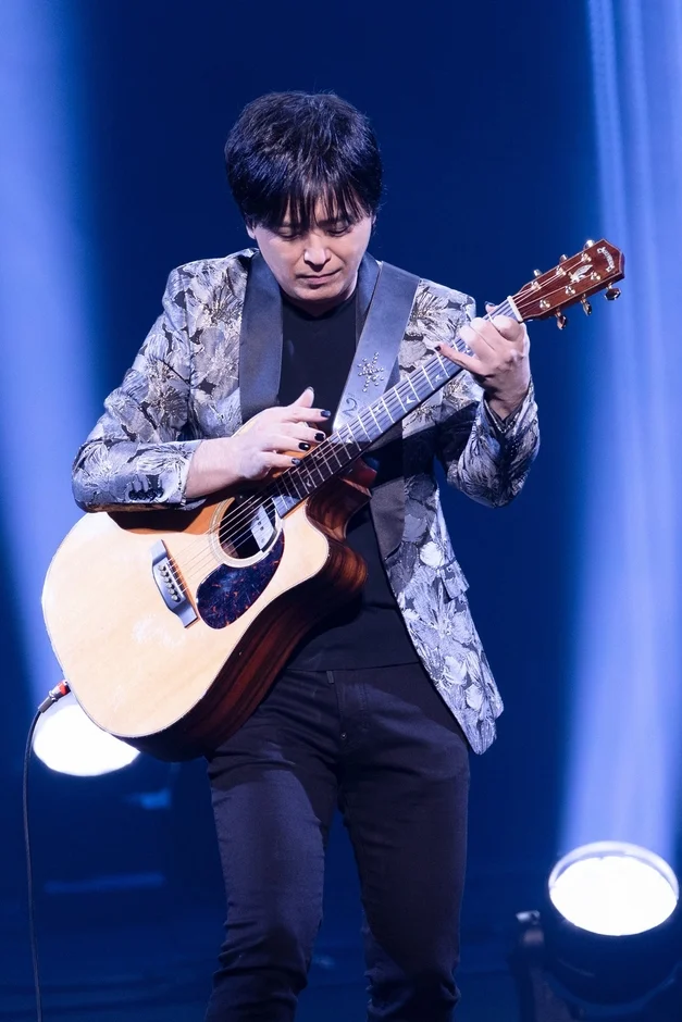 青い照明のステージで、男性ミュージシャンがアコースティックギターを演奏している様子。柄物のジャケットを着用し、黒いマニキュアを施した指で集中してギターを弾いている。