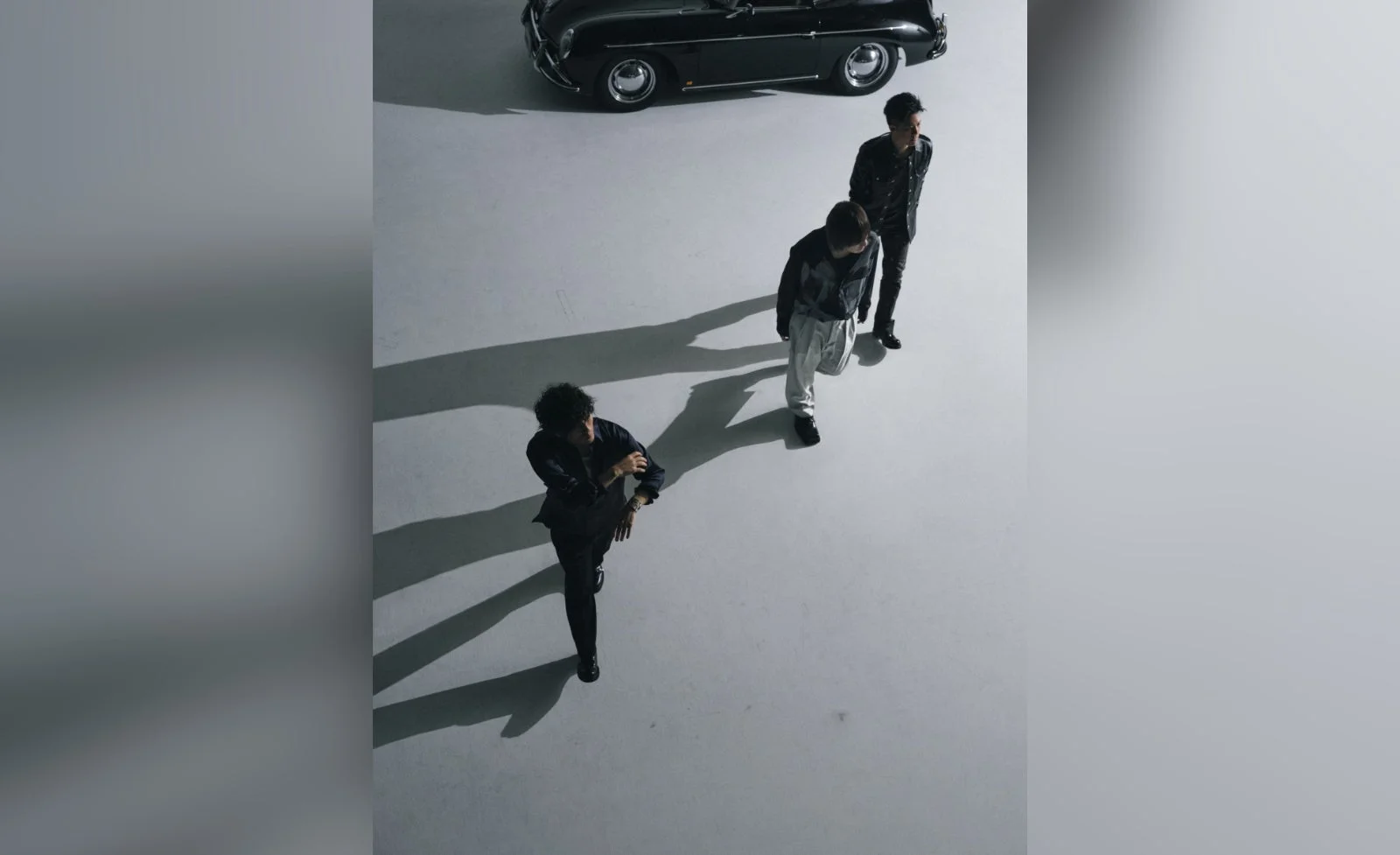 白を基調としたスタジオで、3人の男性が長い影を落としながら一列に歩いている。背景には黒いクラシックカーが停車しており、クールでスタイリッシュな雰囲気を醸し出している。