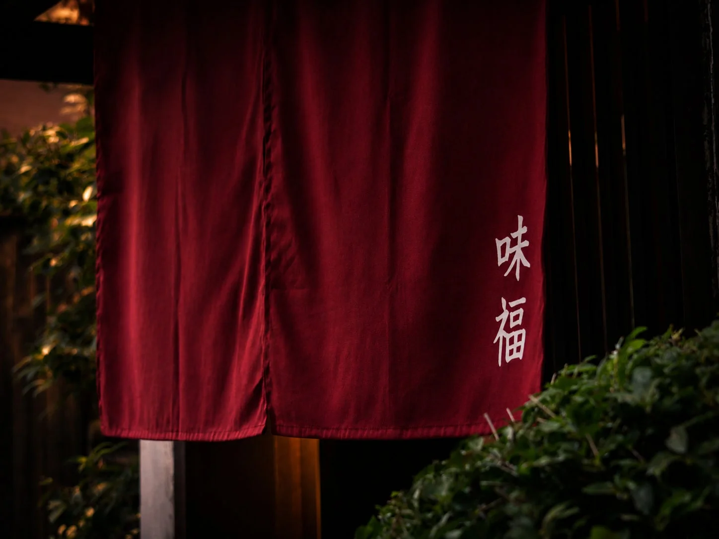 夜の暗がりの中、日本の伝統的な赤い暖簾がかけられており、「味福」という白い文字が縦書きで記されています