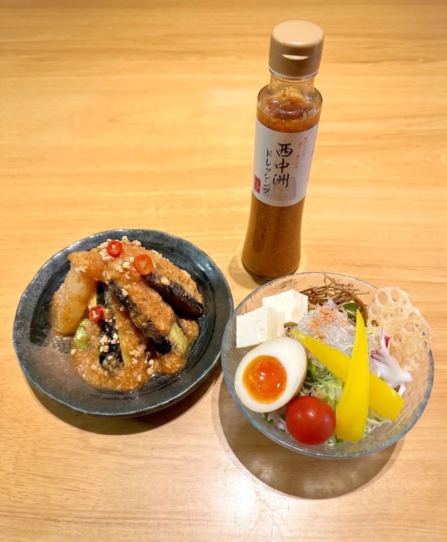 木製のテーブルに、ナス料理と彩り豊かなサラダ、そしてドレッシングのボトルが並んでいます。サラダには半熟卵、豆腐、ミニトマト、パプリカ、海藻、蓮根チップスなどが入っており、ヘルシーでバランスの取れた和食のセットです。