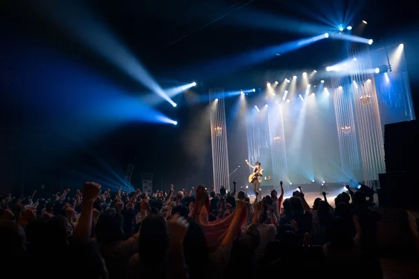 ライブコンサートの様子を捉えた画像です。ステージ上ではギタリストがアコースティックギターを演奏し、青と白のスポットライトがドラマチックに照らしています。手前には多くの観客が腕を上げて熱狂しており、会場全体の高揚感が伝わってきます。