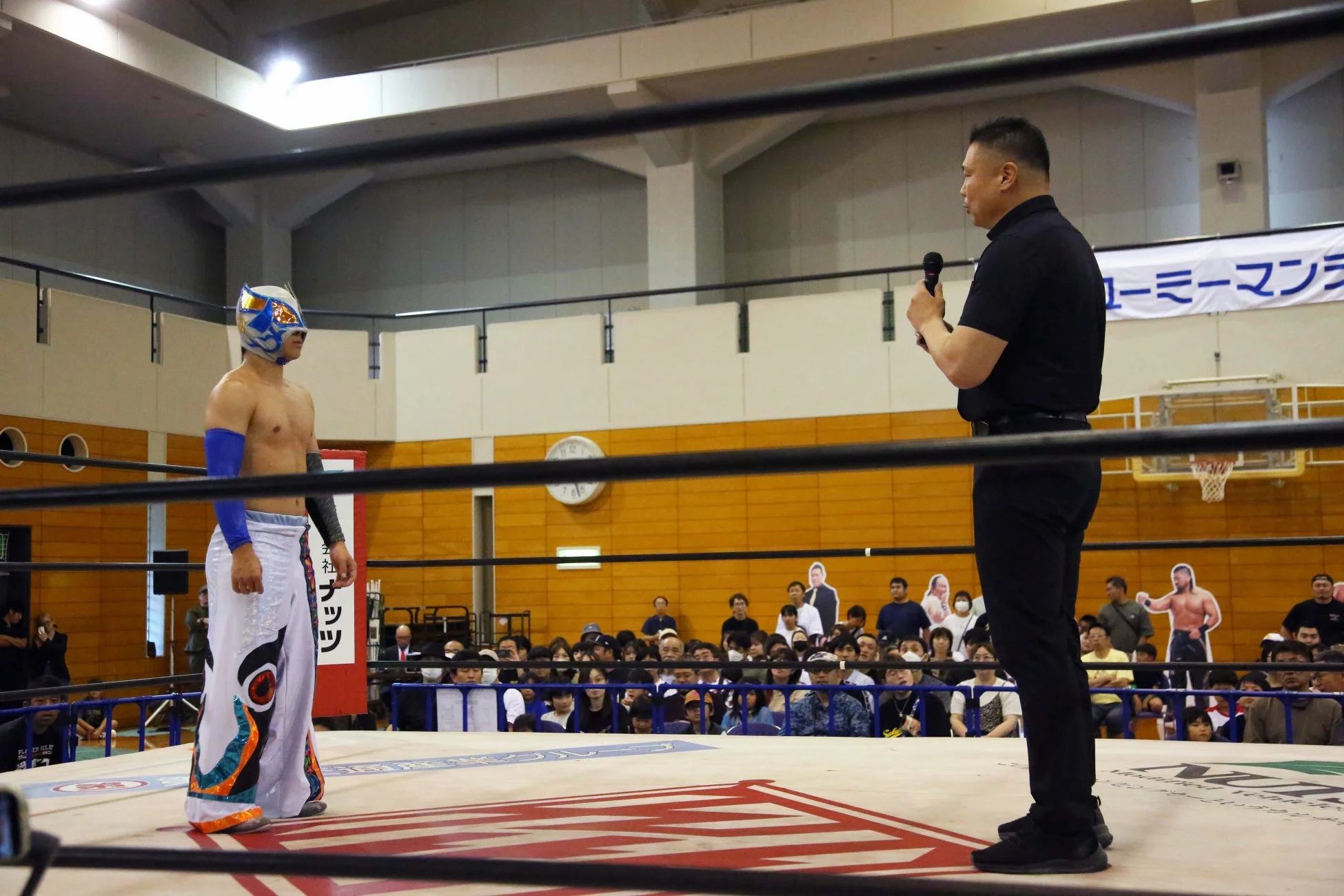 体育館のプロレスリング上で、マスクをかぶったレスラーとマイクを持つ男性が向かい合っている。周囲には多くの観客が見守っている。