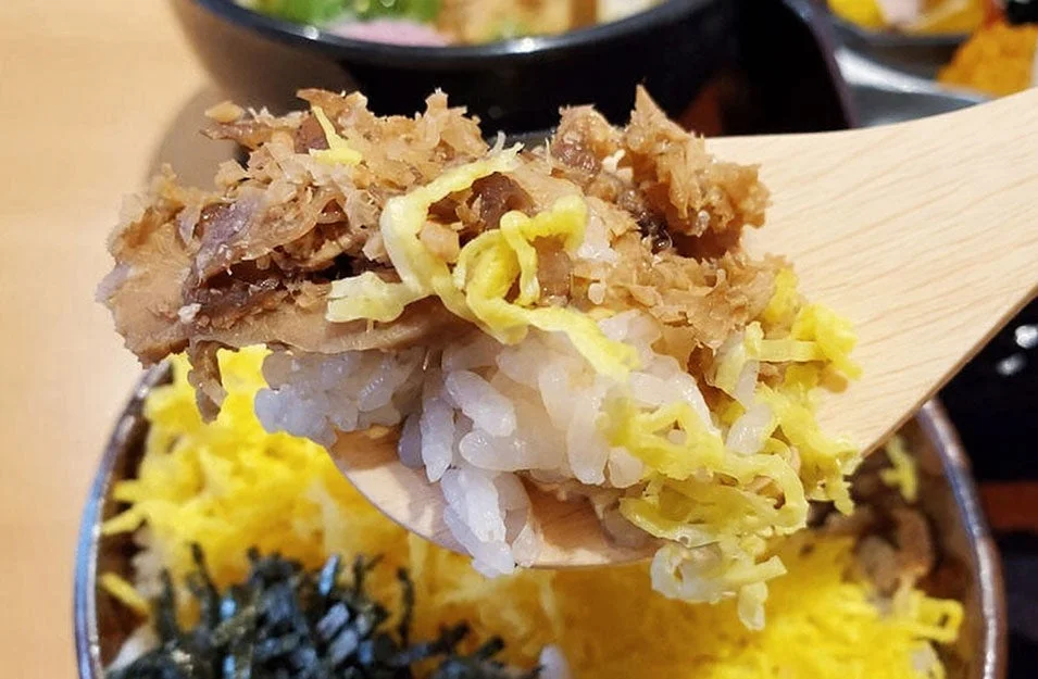 木のスプーンで、ご飯の上に錦糸卵と鶏そぼろが乗った丼物をすくっている様子をクローズアップで捉えた画像です。