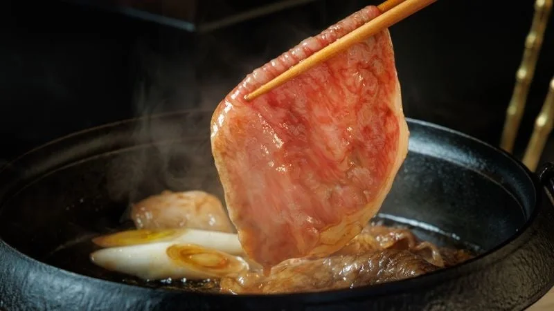 箸で持ち上げられた霜降りの牛肉が、湯気の立つすき焼き鍋の上で輝いています