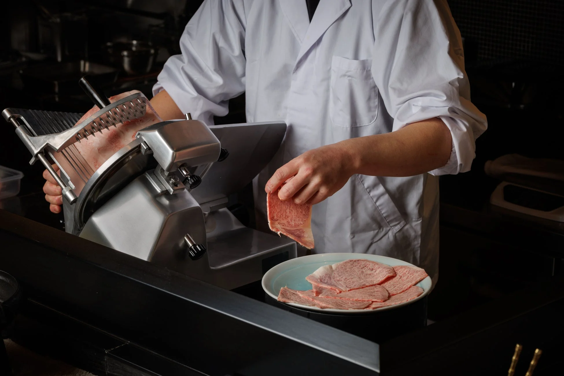 白衣を着た人物が業務用スライサーを使い、霜降りの牛肉を薄くスライスし、皿に盛り付けている様子