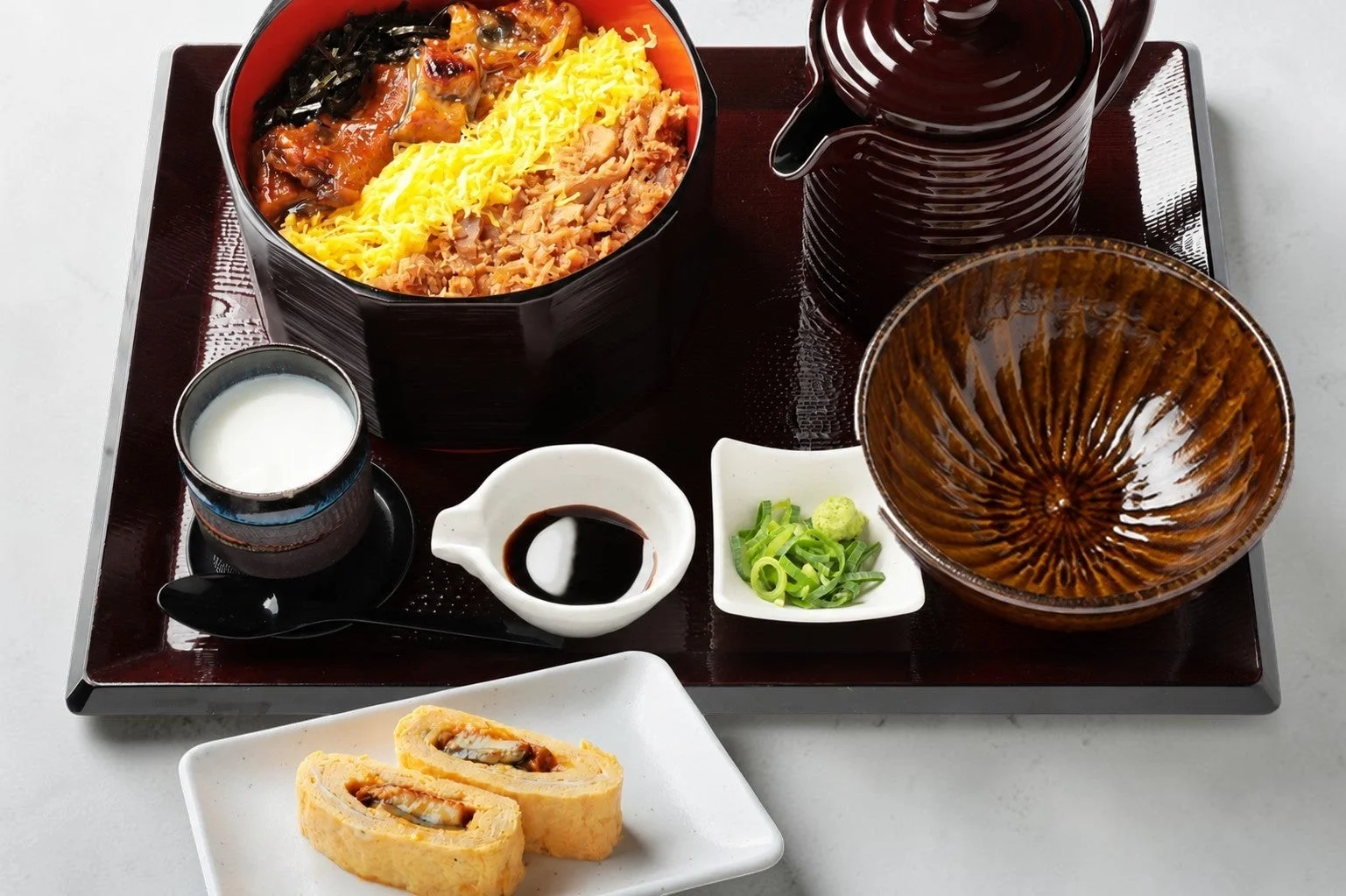 豪華なひつまぶし膳が写っています。大きな丼にはうなぎ、錦糸卵、刻み海苔が盛り付けられ、薬味、出汁が入った急須、う巻き卵、お椀などが朱色の膳に美しく並べられています。