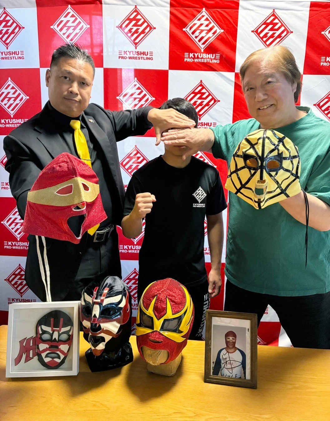 KYUSHU PRO-WRESTLINGのロゴが入った背景の前で、2人の大人と1人の子供がポーズを取る様子。大人はそれぞれレスリングマスクを持ち、子供の目を隠している。テーブルには複数のマスクが飾られており、プロレスに関連するイベントや記念撮影の様子を捉えた一枚。