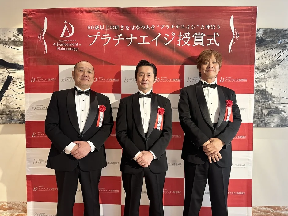 「プラチナエイジ受賞式」の背景の前でポーズをとるタキシード姿の3人の男性