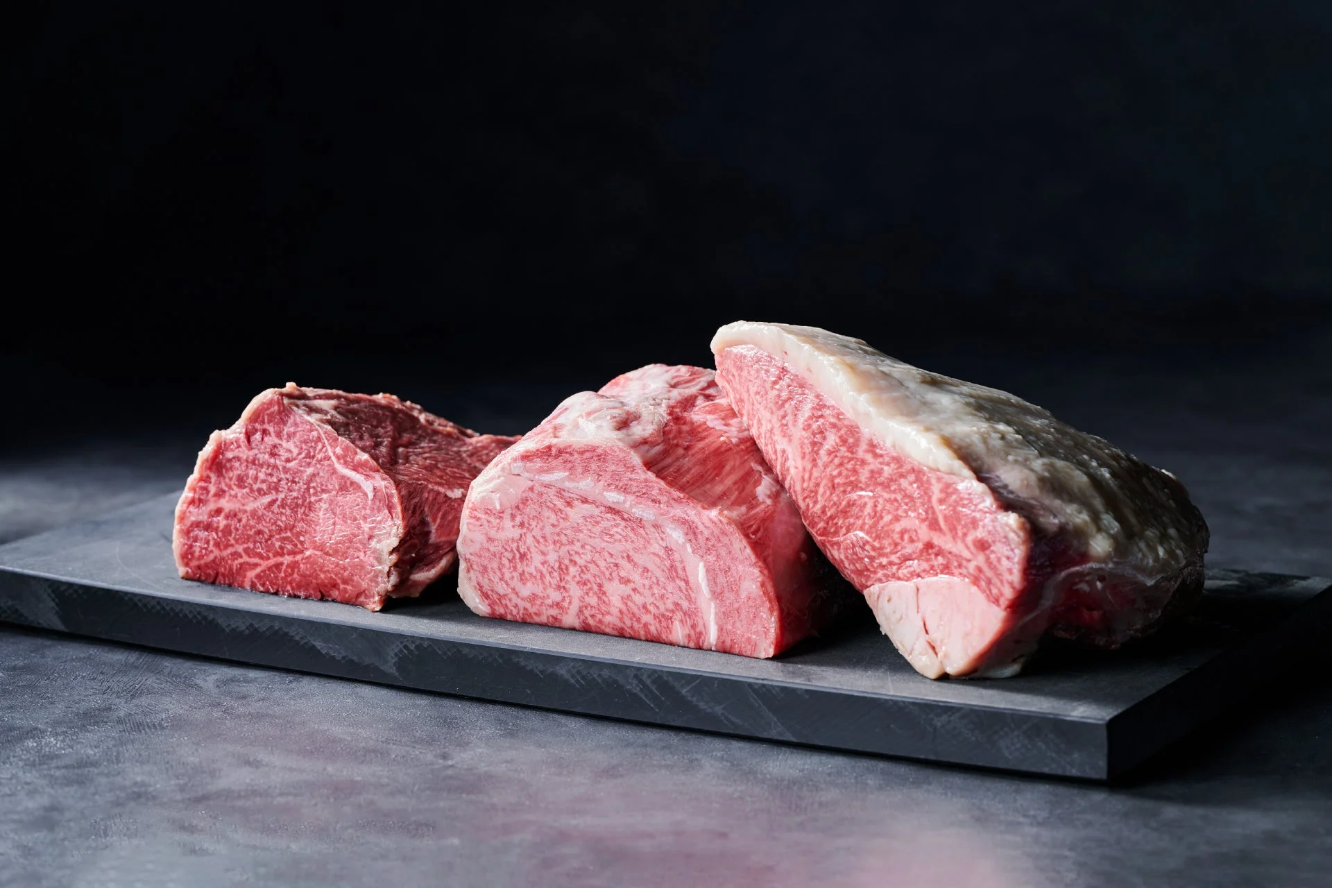 暗い背景と石板の上に、美しい霜降りの高級和牛肉の塊が3つ並べられています