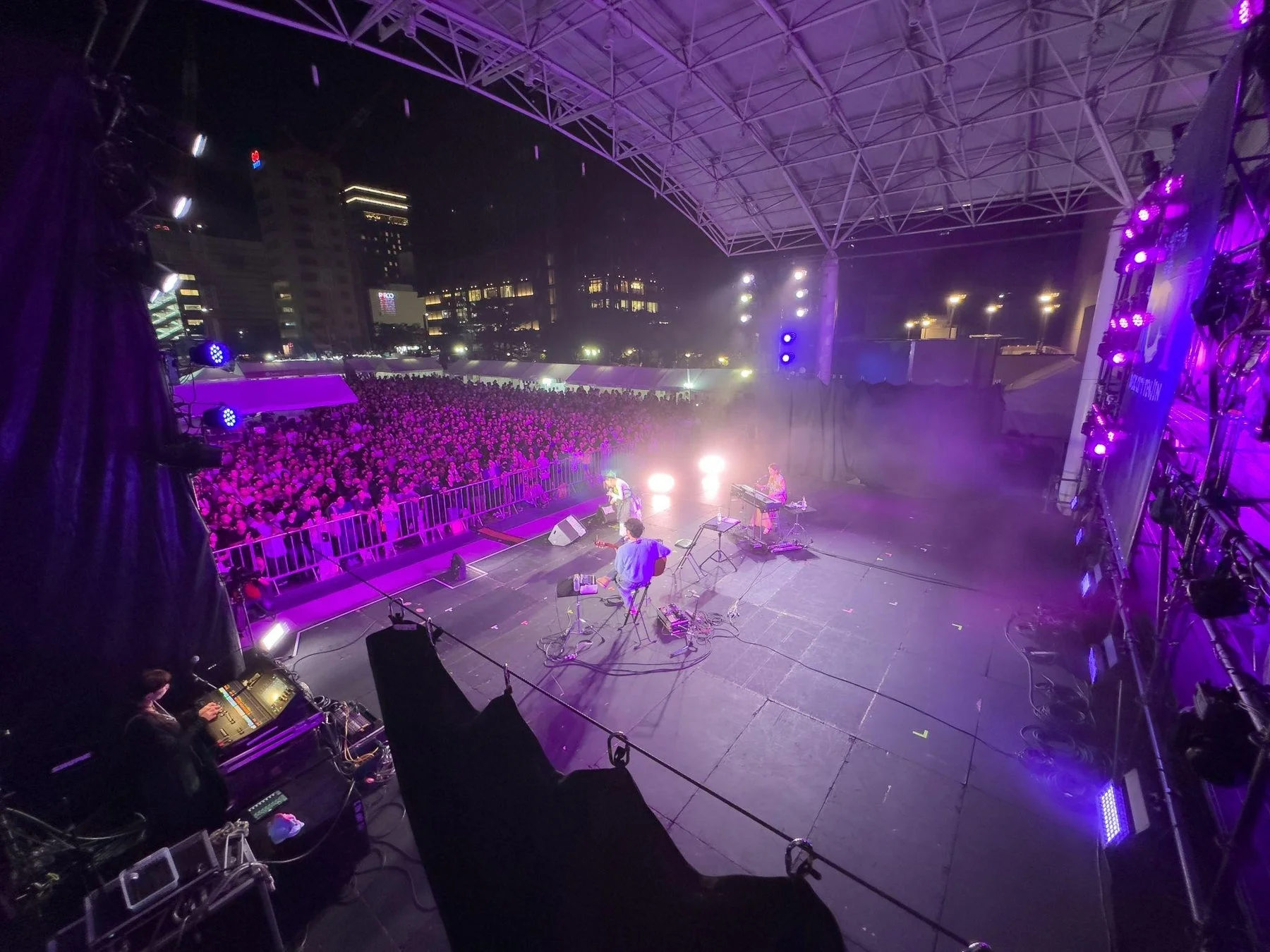 夜の野外ステージでバンドが演奏しており、多数の観客が紫色の照明に照らされているライブイベントの様子