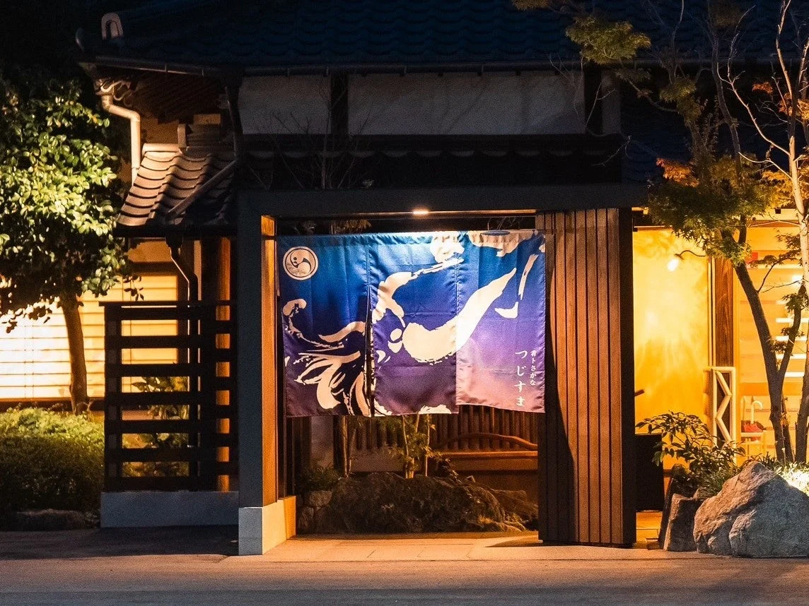 夜の日本家屋の入り口を捉えた画像です。青と紫のグラデーションの暖簾がかかり、「旬のさかな つじしま」と書かれています。温かい光に照らされ、趣のある雰囲気を醸し出しています。