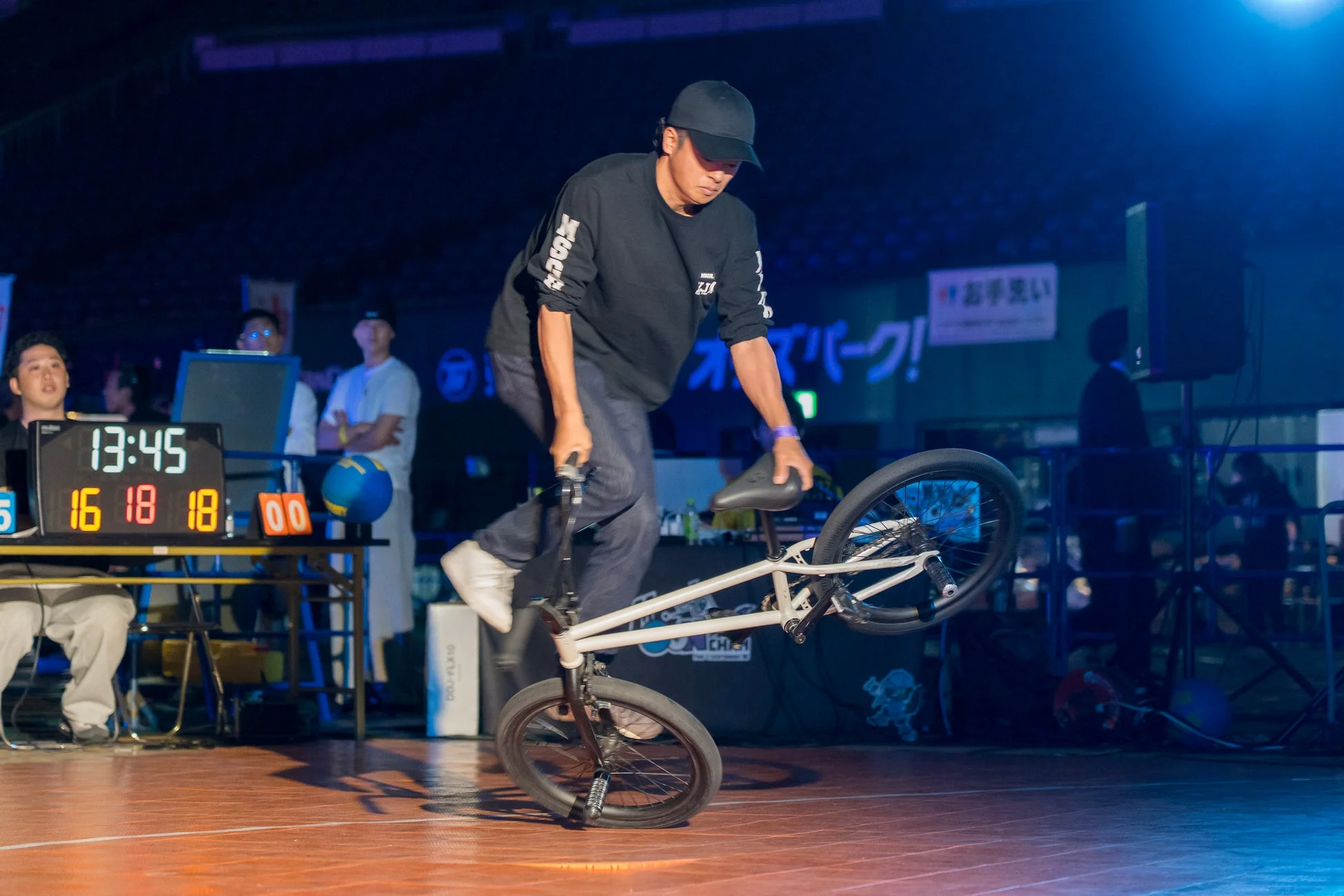 暗い屋内ステージで、男性がBMX自転車でフラットランドのトリックを披露しています。背景にはスコアボードが表示され、競技イベントの様子がうかがえます。