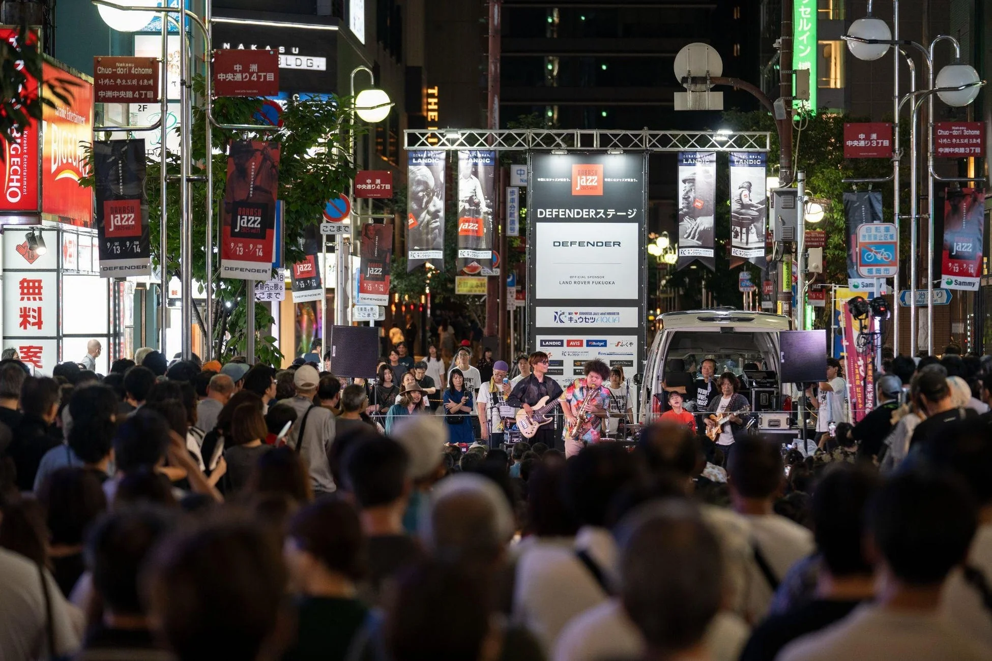 日本の都市の夜景で、中洲ジャズフェスティバルの一環として、野外ステージでバンドがライブ演奏を行っています