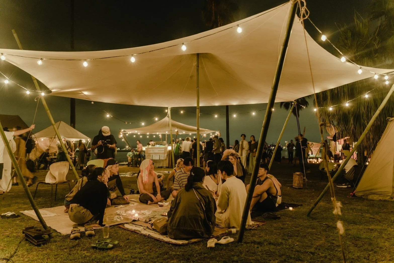 夜の屋外イベントで、人々が大きなテントの下に集まり、地面に座って談笑しています