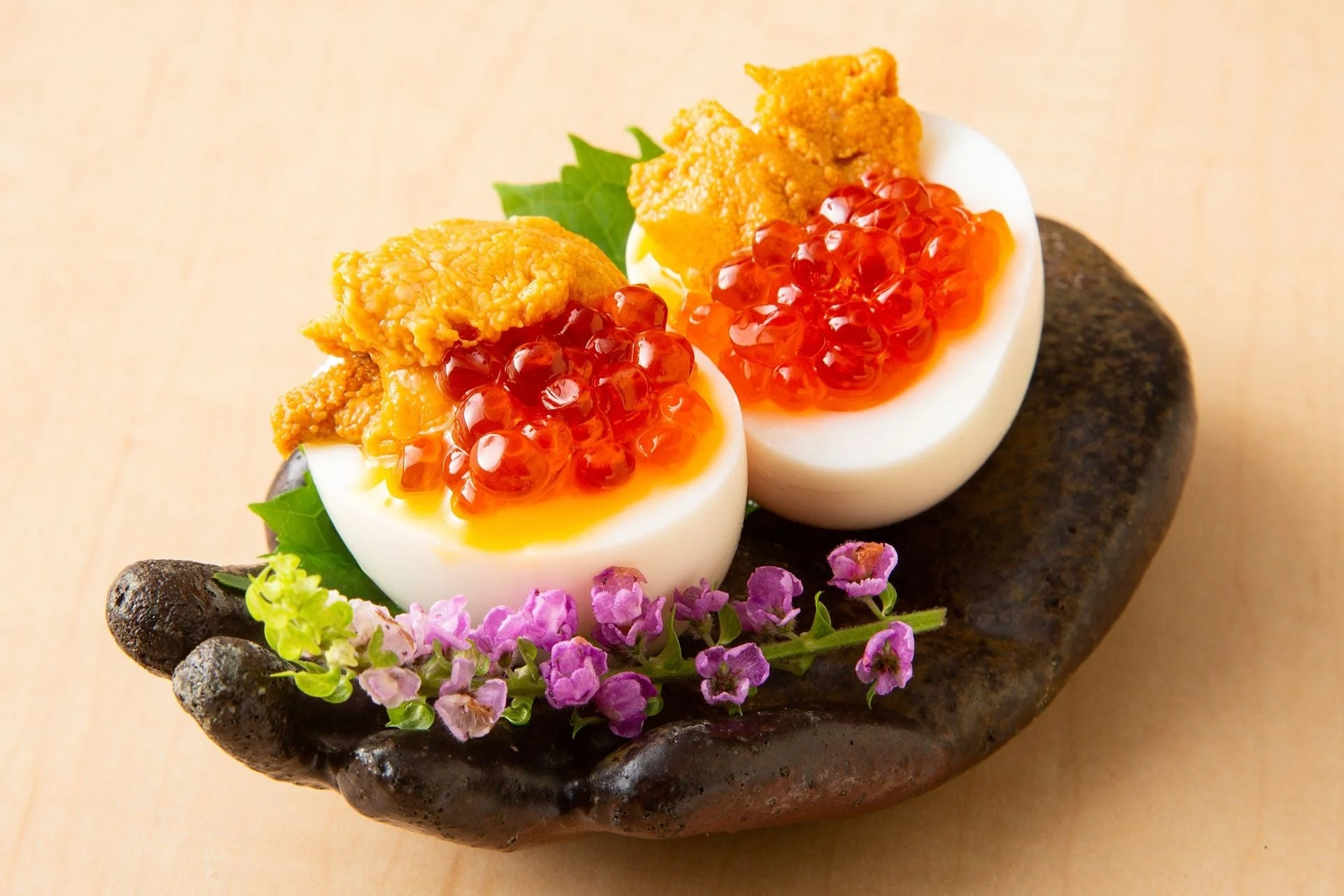 半熟卵の上にいくらとうにが贅沢に盛り付けられた、高級感あふれる和食の一品です。黒い器に添えられた紫色の花が彩りを添えています。