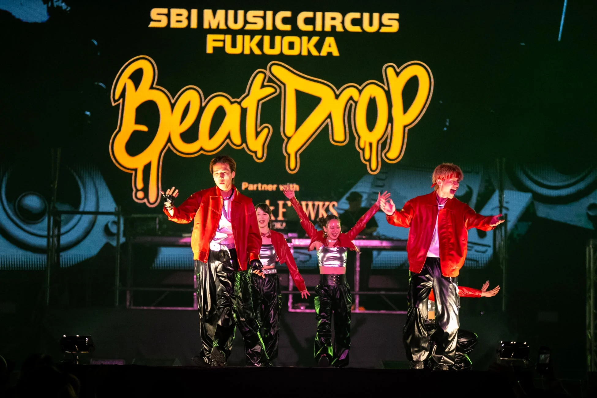 「SBI MUSIC CIRCUS FUKUOKA Beat Drop」のステージで、赤いジャケットを着たパフォーマーたちが躍動しています。巨大なスクリーンにはイベント名が大きく表示されており、活気ある音楽イベントの様子が伝わります。