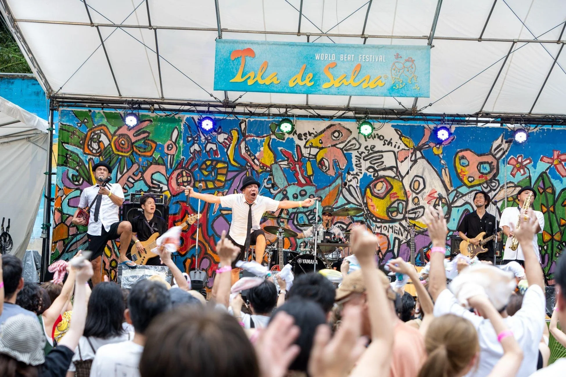 カラフルなアートが描かれたステージでバンドが熱演し、多くの観客が手を挙げて盛り上がっている野外音楽フェスの様子