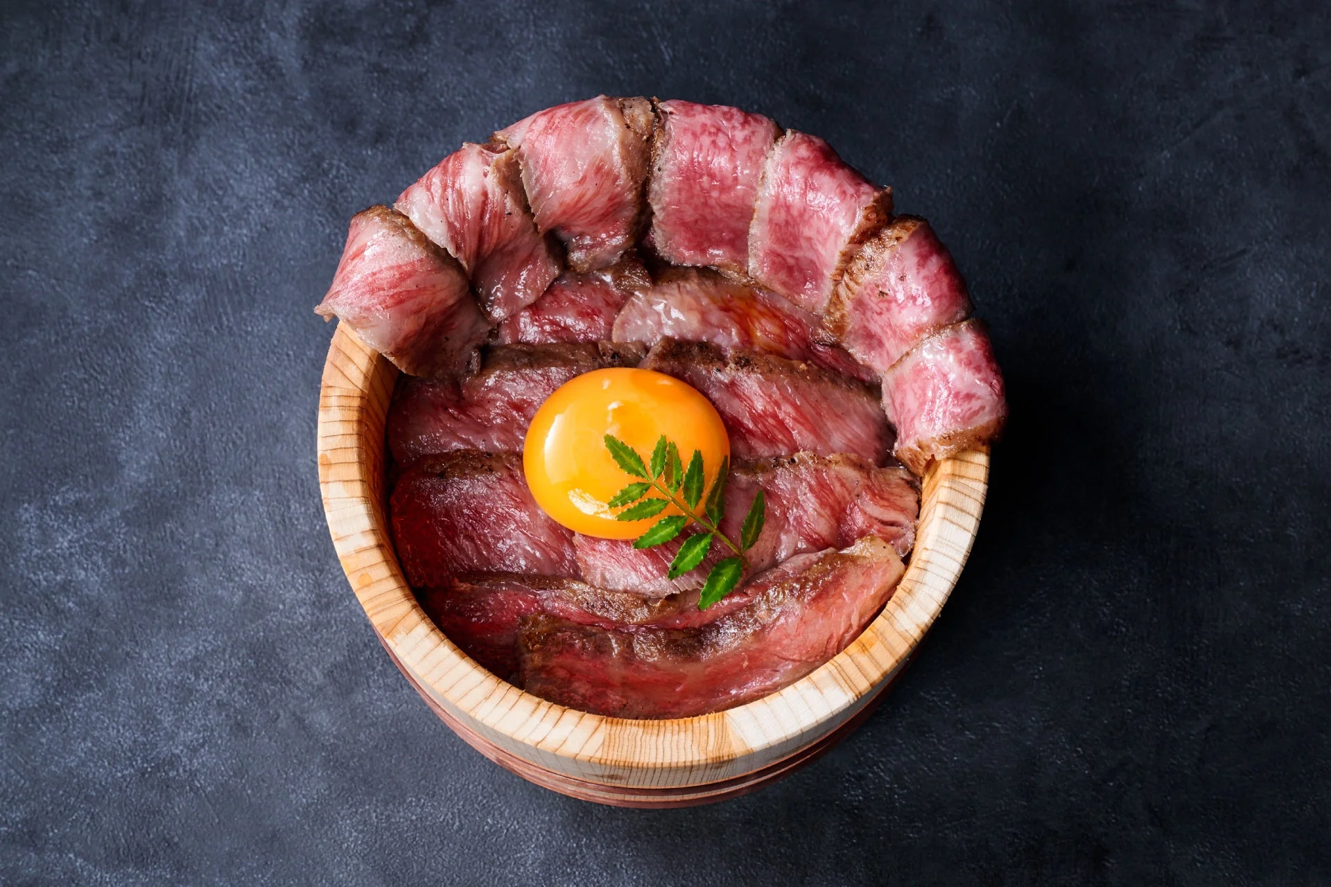 木桶に盛り付けられた薄切りのローストビーフ丼で、中央には鮮やかな卵黄と添えられた緑の葉が美しい一品